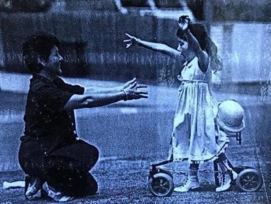 A young girl in a dress with outstretched arms walks toward a kneeling adult, also reaching out, on a grassy area. A hat rests on a small wheeled walker beside the girl.