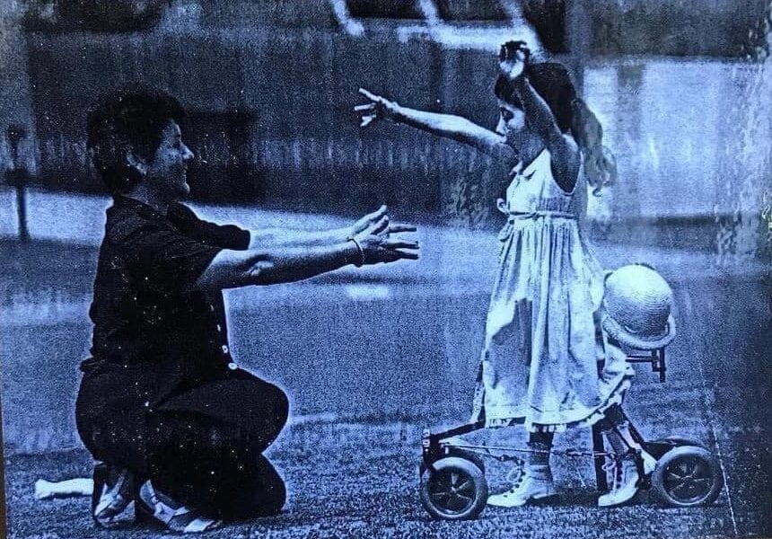 A young girl in a dress with outstretched arms walks toward a kneeling adult, also reaching out, on a grassy area. A hat rests on a small wheeled walker beside the girl.