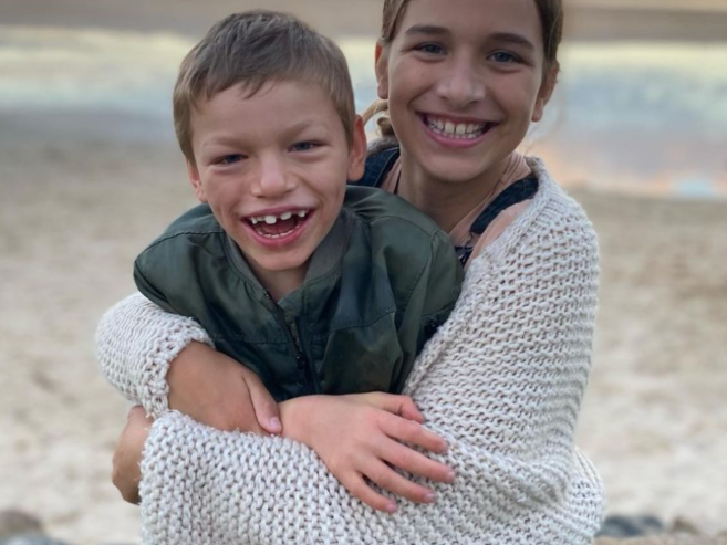 Two smiling children sit together on a beach, with the older child hugging the younger one from behind. The background shows sand, water, and a colorful sky at sunset.