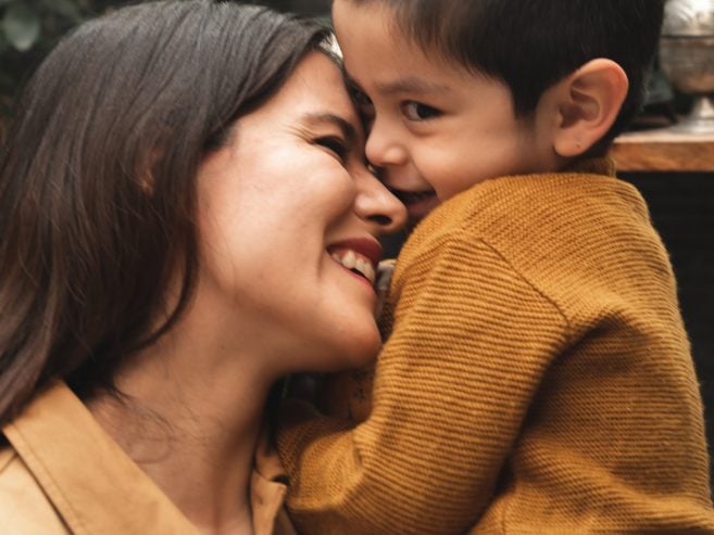 A woman and a young boy, both smiling and wearing tan-brown clothing, cuddle cheek to cheek outdoors with green foliage in the background.