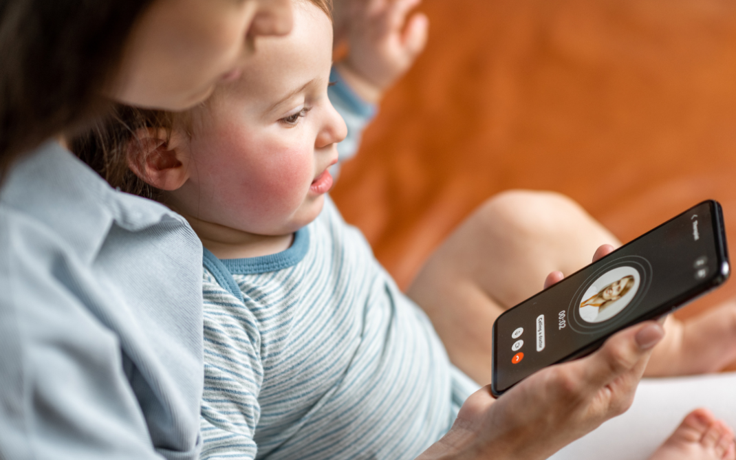 A woman holds a baby on her lap while they look at a smartphone screen showing an incoming video call with a contact named Anna. The baby is focused on the phone, and both are sitting on a bed or couch.