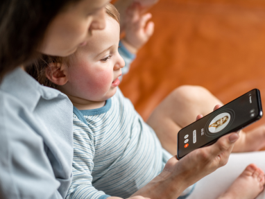 A woman holds a baby on her lap while they look at a smartphone screen showing an incoming video call with a contact named Anna. The baby is focused on the phone, and both are sitting on a bed or couch.