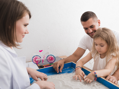 A young girl and her father participate in a sand play therapy session with a female therapist. The girl draws in the sand while the adults watch and support her, and a toy bicycle is visible in the background.