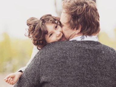 A young child smiles joyfully while being hugged by an adult with light brown hair wearing a gray sweater. The child’s face is pressed against the adult’s shoulder, radiating happiness and affection.