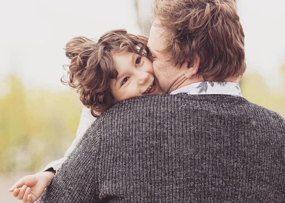 A young child smiles joyfully while being hugged by an adult with light brown hair wearing a gray sweater. The child’s face is pressed against the adult’s shoulder, radiating happiness and affection.