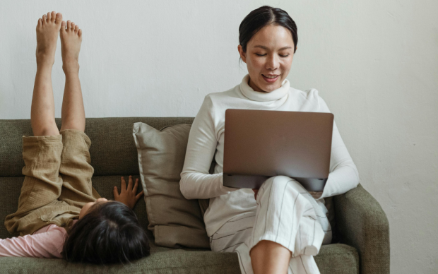 A woman sits on a couch using a laptop while a child lies next to her with legs up against the couch backrest, both appearing relaxed in a home setting.