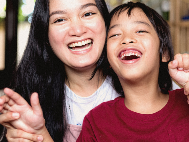 A woman and a child with long dark hair sit close together indoors, smiling and laughing joyfully as they hold hands, creating a warm and happy atmosphere.