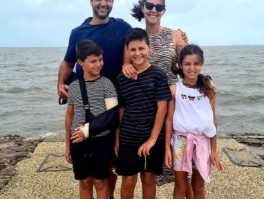 A smiling family of five poses by the seaside. Two adults stand behind three children; one boy has his arm in a sling. The sky is cloudy and the sea is calm in the background.