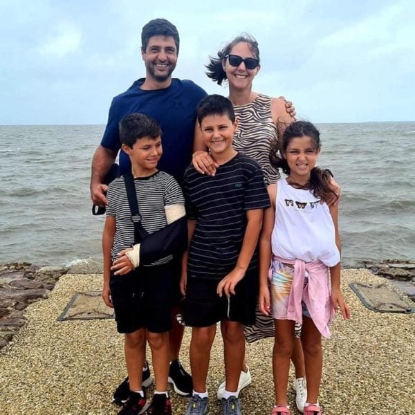 A smiling family of five poses by the seaside. Two adults stand behind three children; one boy has his arm in a sling. The sky is cloudy and the sea is calm in the background.