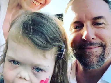 A smiling man and woman take a selfie outdoors with a young girl who has a pink heart painted on her cheek. The girl is wearing a denim jacket and appears happy. The background shows a clear, sunny sky.