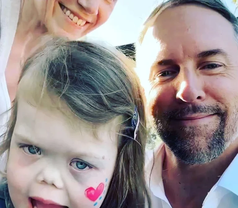 A smiling man and woman take a selfie outdoors with a young girl who has a pink heart painted on her cheek. The girl is wearing a denim jacket and appears happy. The background shows a clear, sunny sky.