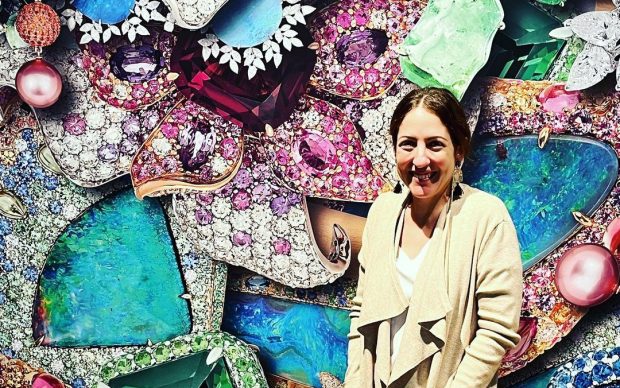 A woman wearing a beige cardigan stands smiling in front of a colorful, intricate wall composed of large gemstone and jewelry designs.
