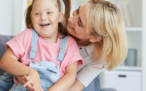 A woman with blonde hair is sitting on a couch, hugging and smiling at a young girl with Down syndrome who is laughing and wearing a pink shirt with denim overalls.