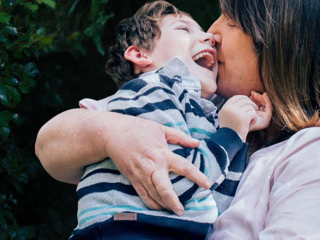A woman smiles and cuddles a laughing young boy in her arms, both enjoying a joyful moment outdoors with green foliage in the background.