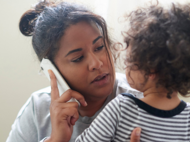 A woman holds a young child while talking on the phone, looking concerned. The child faces her, both indoors with a light background.
