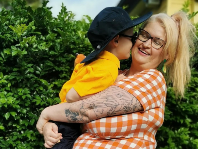 A smiling woman in glasses and an orange checkered shirt holds a young child who is whispering in her ear. They are outdoors in front of a green hedge on a sunny day.