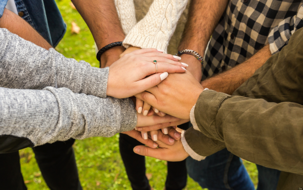 A group of people standing in a circle outdoors place their hands together in the center, showing unity and teamwork. The grass beneath them is green, and only their arms and torsos are visible.