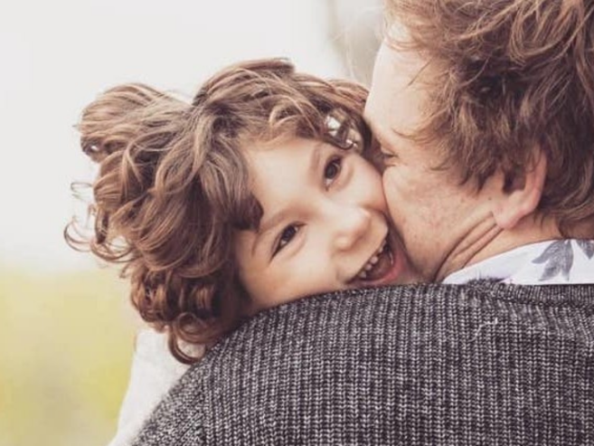 A young child with curly hair smiles happily while being embraced by an adult from behind, outdoors on a bright day. The childs head rests on the adults shoulder.