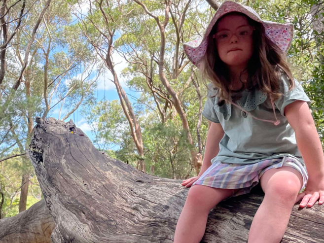 A young girl wearing glasses and a sunhat sits on a large fallen tree trunk in a sunlit forest, surrounded by tall trees and greenery under a partly cloudy sky.