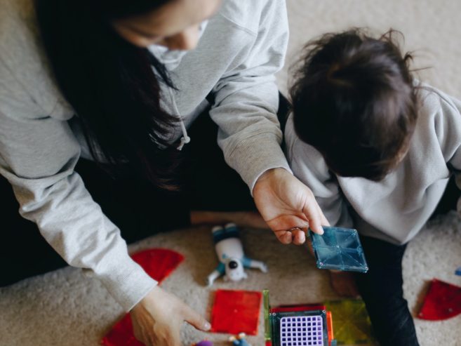 A woman and a young child sit on a carpeted floor, playing with colorful magnetic tiles and toys. The woman holds a blue tile while the child looks at the pieces, both engaging in a hands-on activity.