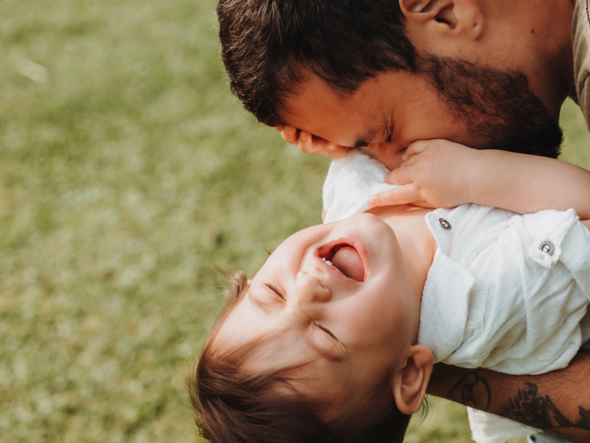 A bearded man with tattoos gently tickles a laughing baby, who is wearing a white shirt, while playing together on green grass outdoors.