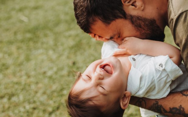 A bearded man with tattoos gently tickles a laughing baby, who is wearing a white shirt, while playing together on green grass outdoors.