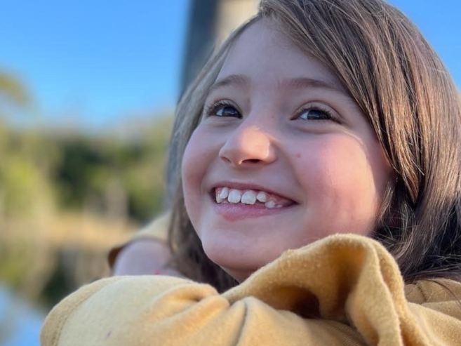 A young girl with long brown hair and a yellow sweater smiles brightly while sitting by a calm lake, with trees and a blue sky in the background.