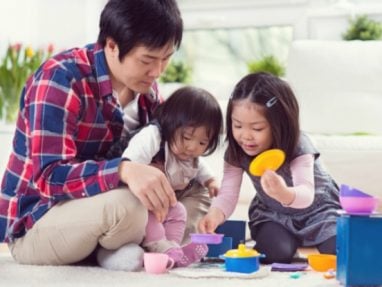 A father sits on the floor with his two young daughters, playing with colorful toy dishes and blocks in a bright, cozy living room. The children are focused and smiling as they play together.
