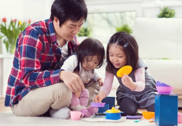 A father sits on the floor with his two young daughters, playing with colorful toy dishes and blocks in a bright, cozy living room. The children are focused and smiling as they play together.