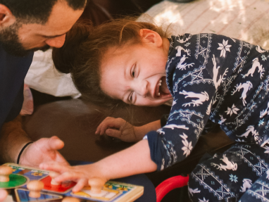 A man helps a smiling young girl in patterned pajamas play with a wooden toy while she sits in a wheelchair.