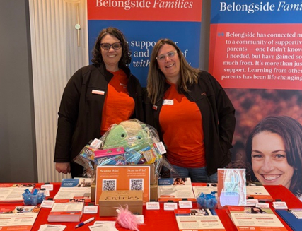 Two women stand behind a display table with brochures, QR codes, and colorful items. A basket with stuffed toys sits on the table. Behind them are banners for Belongside Families with information and a smiling childs photo.