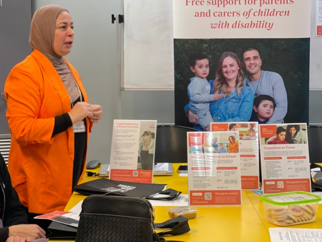 A woman in an orange jacket speaks at a table with brochures about support for parents and carers of children with disability. A large poster behind her shows a smiling family, including two children.