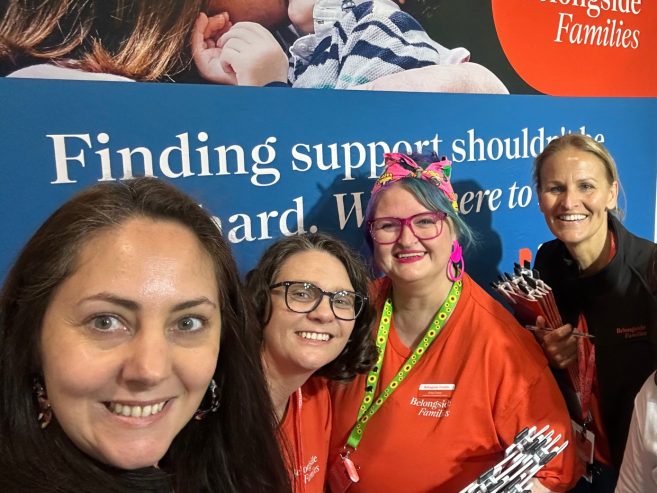 Four smiling women, three wearing red Belongside Families shirts, stand together at an event booth in front of a banner that reads, Finding support should be easy. Were here to help.
