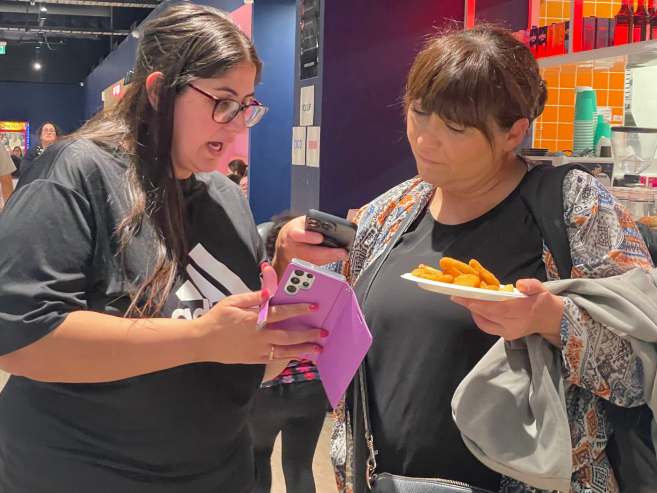 Two women stand indoors. One is holding a phone with a purple case and showing the screen to the other, who is holding a plate of food and a jacket. Both appear focused on the phone. People and shelves are in the background.