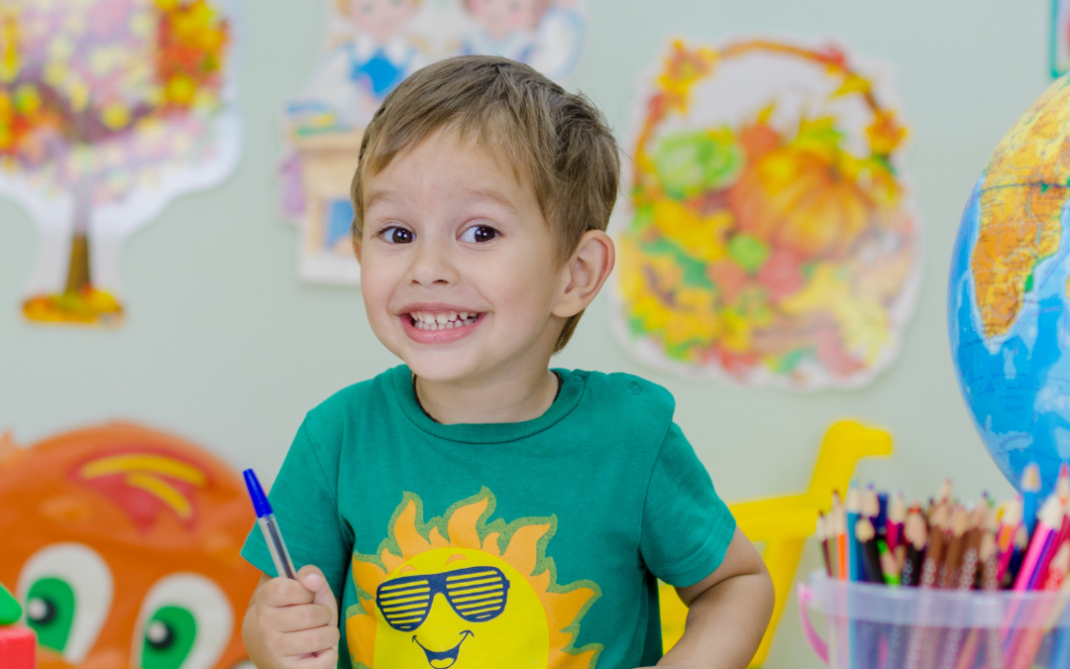 A young boy in a green shirt with a smiling sun design is sitting at a table, smiling and drawing in a book. Colorful art supplies, a globe, and drawings of autumn trees are visible in the background.