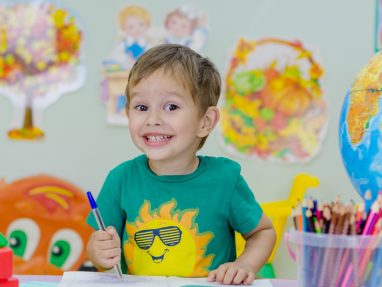 A young boy in a green shirt with a smiling sun design is sitting at a table, smiling and drawing in a book. Colorful art supplies, a globe, and drawings of autumn trees are visible in the background.