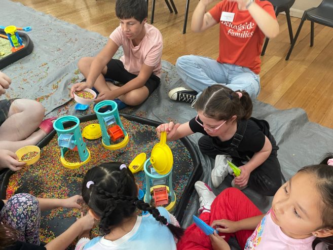 Children sit on the floor around sensory play trays filled with colorful rice, scooping and pouring it through plastic funnels and wheels. They are focused on playing and interacting together in a bright indoor setting.