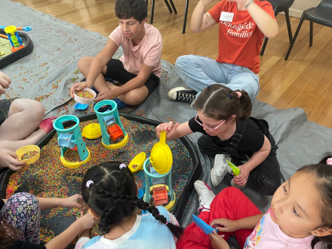 Children sit on the floor around sensory play trays filled with colorful rice, scooping and pouring it through plastic funnels and wheels. They are focused on playing and interacting together in a bright indoor setting.