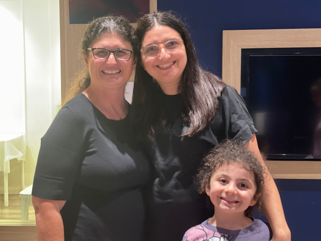 Three people, two adults and one child, smile while posing together indoors. The adults have dark hair and wear black tops, and the child has curly hair and wears a colorful shirt. A TV and blue wall are in the background.