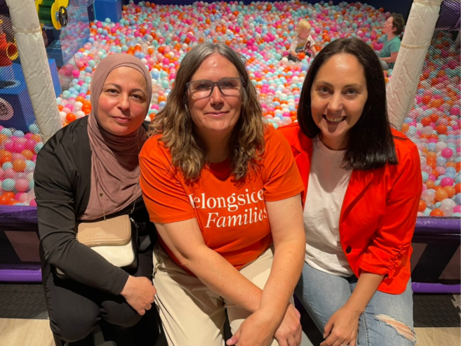Three women sit closely together in front of a colorful ball pit, smiling at the camera. The woman in the center wears glasses and an orange shirt that reads Belonging is for Families.