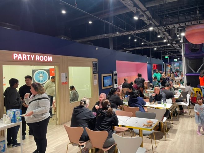 A lively indoor party room with groups of people sitting at tables, eating, and chatting. Some people stand near a door labeled Party Room while children play in the background. The atmosphere is bright and social.