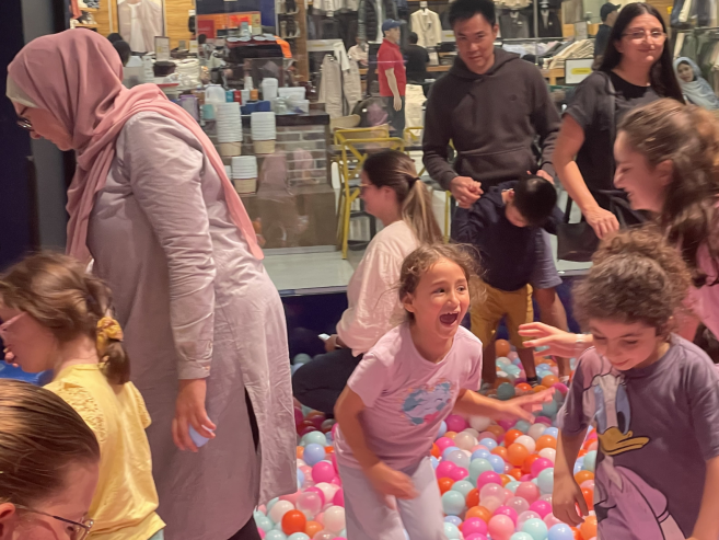 Children play and laugh together in a colorful ball pit, surrounded by adults. The scene is lively and joyful, with a clothing store visible in the background through glass windows.