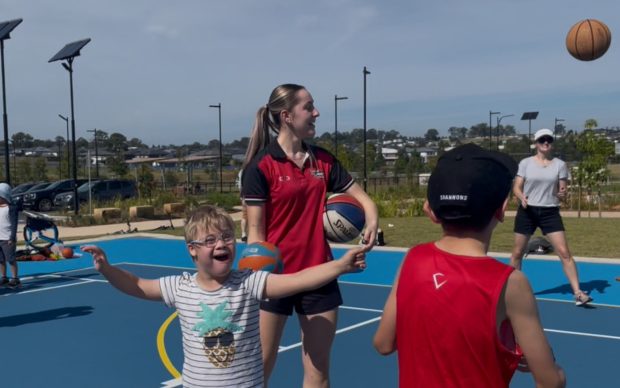 A smiling child with glasses gestures excitedly on an outdoor basketball court, while a woman in a red shirt holds a basketball. Other kids and adults play in the background under a sunny sky.