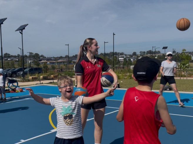 A smiling child with glasses gestures excitedly on an outdoor basketball court, while a woman in a red shirt holds a basketball. Other kids and adults play in the background under a sunny sky.