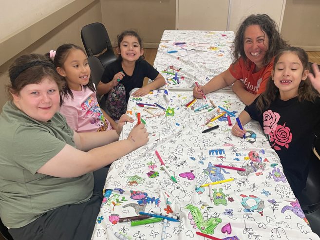 Five people, including four children and an adult, sit around a table covered with a coloring sheet. They are smiling and holding markers, engaged in coloring together. The table is decorated with colorful drawings.
