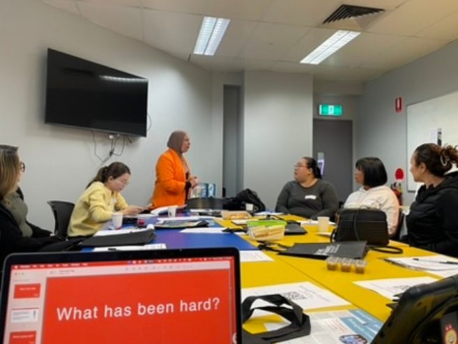 A group of people sit around a conference table with papers and laptops, listening to a person in an orange blazer speaking. A presentation slide on a laptop reads, “What has been hard?”.