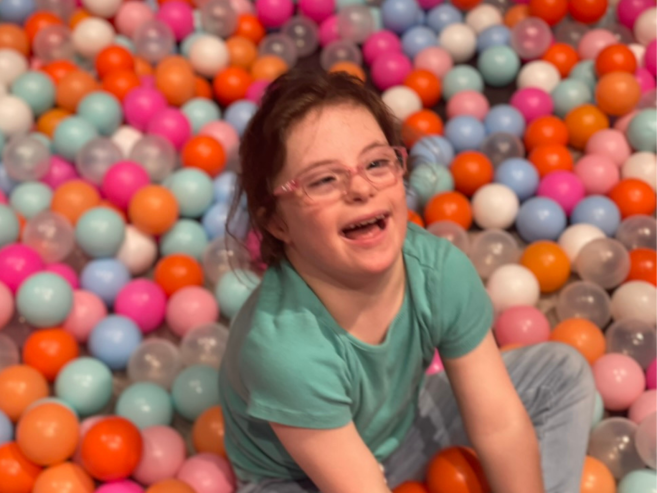 A smiling child with glasses sits in a colorful ball pit filled with orange, pink, white, and blue balls. She is wearing a turquoise shirt and light blue pants.