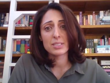 A woman with shoulder-length brown hair and a green shirt sits in front of a bookshelf filled with books, looking slightly concerned. A white ladder leans against the shelves in the background.