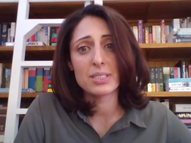 A woman with shoulder-length brown hair and a green shirt sits in front of a bookshelf filled with books, looking slightly concerned. A white ladder leans against the shelves in the background.