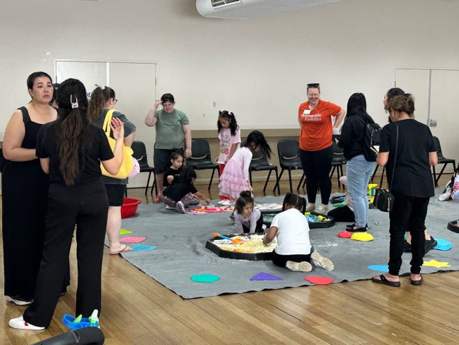 A group of adults and children participate in indoor activities on a large gray mat, with colorful items scattered around. Some kids sit and play while adults stand nearby watching or talking.